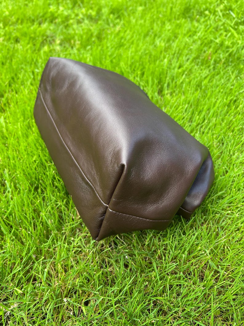 Puede incluir: Un estuche de cuero marr&oacute;n oscuro con un acabado liso y mate y un dise&ntilde;o estructurado y plegado. El art&iacute;culo presenta l&iacute;neas limpias y una textura suave y flexible, mostrando un color profundo y uniforme en toda su superficie exterior.