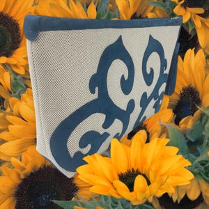 Puede incluir: Una cartera de mano de tela beige y azul con un diseño decorativo. La cartera tiene una cremallera y un borla.