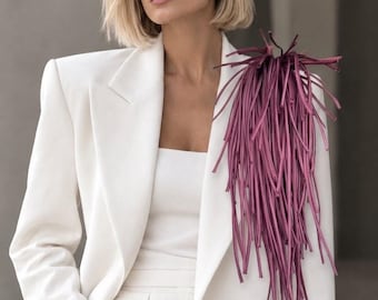 May include: A crisp white blazer featuring structured shoulders and a long, cascading fringe detail in a deep plum colour attached to the left shoulder. The garment is worn over a matching white top, creating a clean, monochromatic look.