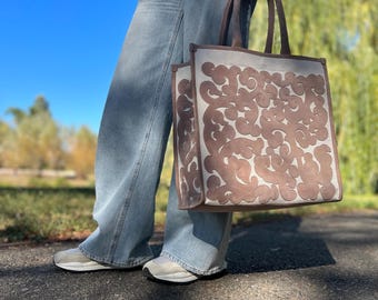 May include: A large, off-white tote bag with brown leather cutouts in a floral pattern. The bag has two brown leather handles.