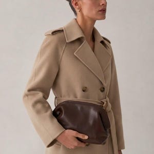 Puede incluir: Un abrigo de lana beige de doble botonadura con cuello de solapa, charreteras en los hombros y cintur&oacute;n a juego, combinado con un bolso de mano de cuero marr&oacute;n oscuro con cierre de marco redondeado y silueta suave y estructurada.