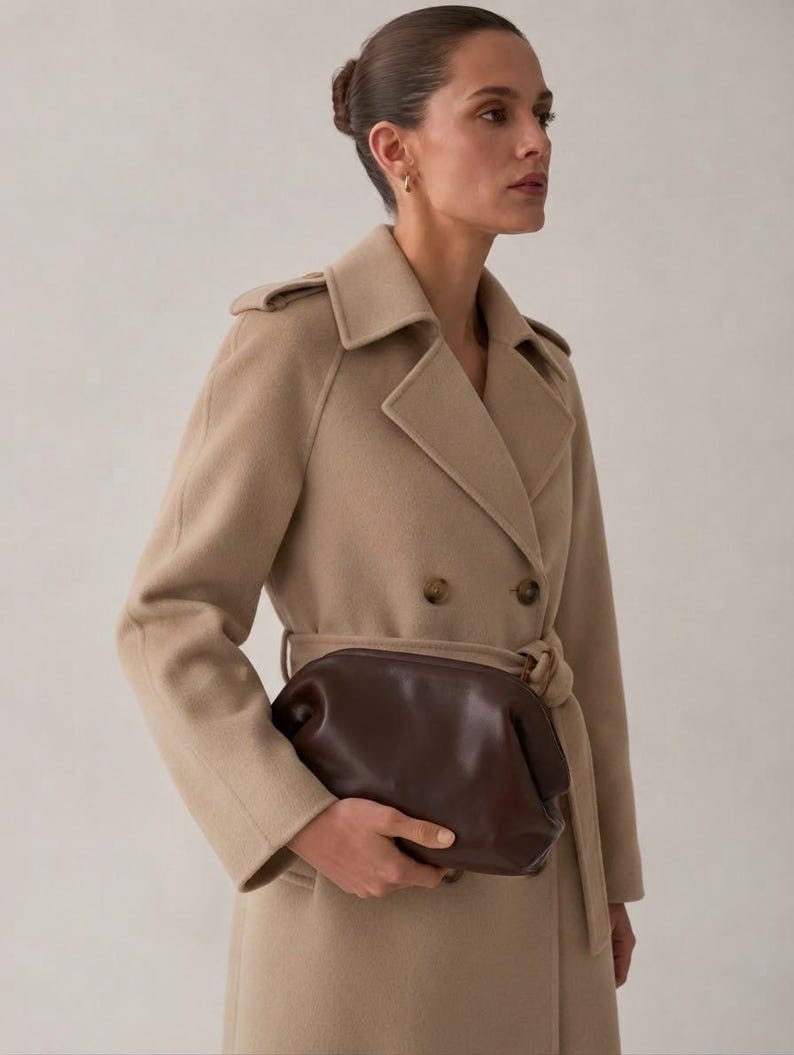 Puede incluir: Un abrigo de lana beige de doble botonadura con cuello de solapa, charreteras en los hombros y cintur&oacute;n a juego, combinado con un bolso de mano de cuero marr&oacute;n oscuro con cierre de marco redondeado y silueta suave y estructurada.