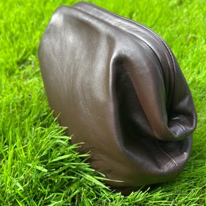 Puede incluir: Un bolso de mano tipo sobre de cuero marr&oacute;n oscuro con una silueta suave, redondeada y sin estructura. El bolso presenta un acabado liso y mate, con un dise&ntilde;o de cierre superior fruncido que crea pliegues naturales en el cuero.
