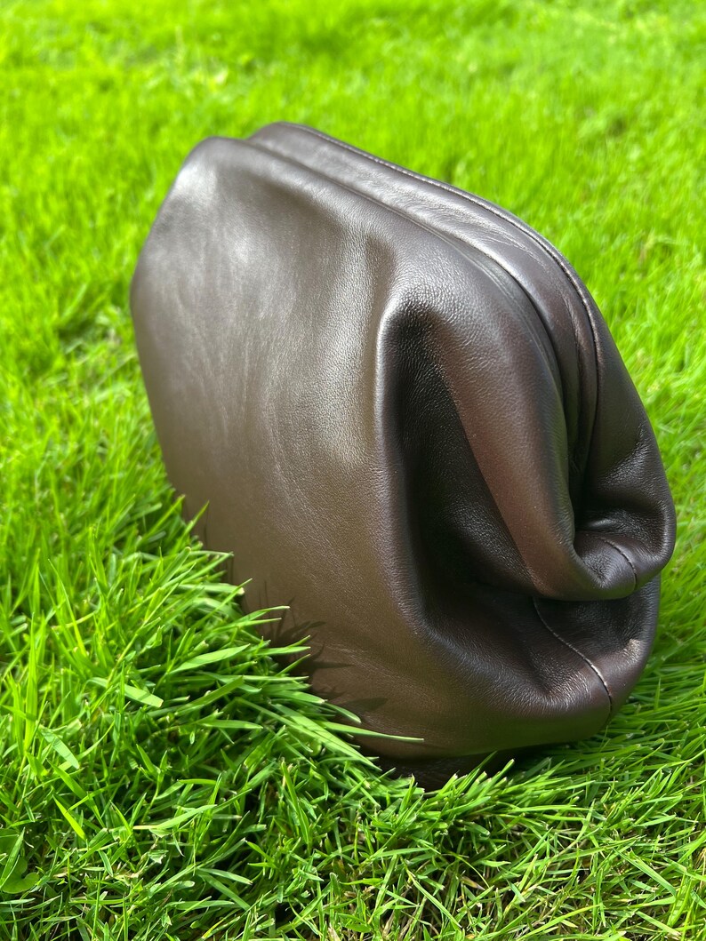 Puede incluir: Un bolso de mano tipo sobre de cuero marr&oacute;n oscuro con una silueta suave, redondeada y sin estructura. El bolso presenta un acabado liso y mate, con un dise&ntilde;o de cierre superior fruncido que crea pliegues naturales en el cuero.