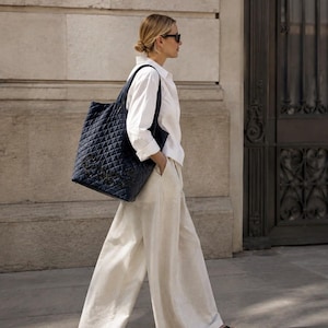 May include: A navy blue quilted tote bag with a diamond pattern and a dark logo on the front, paired with a crisp white long-sleeved button-down shirt, wide-leg off-white trousers, and black sandals. The ensemble is completed with dark sunglasses.