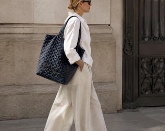May include: A navy blue quilted tote bag with a diamond pattern and a dark logo on the front, paired with a crisp white long-sleeved button-down shirt, wide-leg off-white trousers, and black sandals. The ensemble is completed with dark sunglasses.