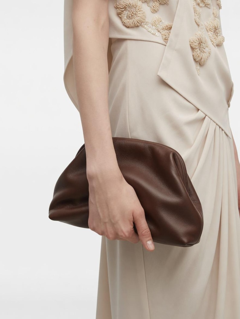 Puede incluir: Un bolso de mano tipo sobre en cuero marr&oacute;n oscuro con una silueta suave, redondeada y tipo bolsa, con un cierre de marco fruncido. El bolso se sostiene contra un vestido sin mangas beige claro con adornos texturizados de flores.
