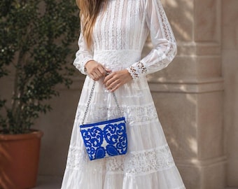 May include: A white, long-sleeved dress with lace inserts and a high neckline. The dress features a tiered skirt and is paired with a small blue and white embroidered purse with a silver chain.