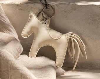 May include: A cream-coloured leather horse-shaped keyring featuring visible stitching along the edges, a saddle detail on the back, a fringed mane, and a long fringed tail. The horse is attached to a silver-toned metal split ring and a lobster clasp.