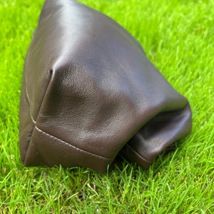 Puede incluir: Un estuche de color marr&oacute;n oscuro hecho de un material liso y flexible con un brillo suave. El art&iacute;culo presenta un dise&ntilde;o estructurado y plegado con costuras visibles a lo largo del borde inferior, creando una forma redondeada y voluminosa.