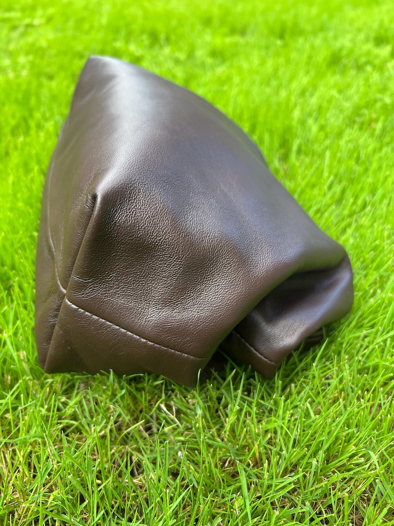 Puede incluir: Un estuche de color marr&oacute;n oscuro hecho de un material liso y flexible con un brillo suave. El art&iacute;culo presenta un dise&ntilde;o estructurado y plegado con costuras visibles a lo largo del borde inferior, creando una forma redondeada y voluminosa.