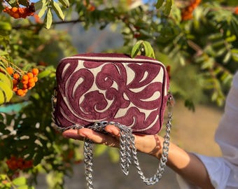 May include: A small burgundy velvet bag with a floral design and a silver chain strap. The bag is held in a hand against a background of green leaves and orange berries.