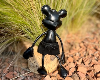 May include: A black leather-like mouse figurine with rounded ears, thin limbs, and black fabric shorts. The figure features visible stitching along the edges of the head and body, with a smooth, dark finish on the head, arms, and feet.