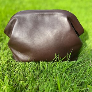Puede incluir: Un bolso de mano tipo sobre de cuero marr&oacute;n oscuro con un acabado liso y mate y una estructura redondeada. El bolso presenta una abertura superior ancha y curva y un cuerpo suave y flexible que se estrecha ligeramente hacia la parte inferior.
