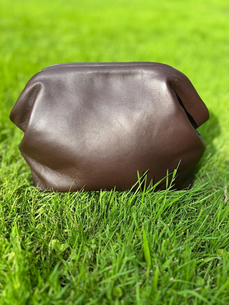 Puede incluir: Un bolso de mano tipo sobre de cuero marr&oacute;n oscuro con un acabado liso y mate y una estructura redondeada. El bolso presenta una abertura superior ancha y curva y un cuerpo suave y flexible que se estrecha ligeramente hacia la parte inferior.