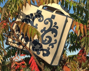 May include: A white canvas tote bag with a blue and white floral design. The bag is hanging from a tree branch with red leaves.