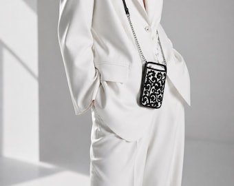 May include: A black and white phone case with an ornate floral design is worn over a white suit. The case has a black strap with a silver chain. The suit consists of a blazer and wide-leg trousers.