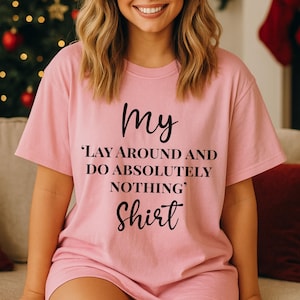 May include: A light pink short-sleeved t-shirt featuring black text that reads: My 'LAY AROUND AND DO ABSOLUTELY NOTHING' Shirt. The garment has a crew neckline and a relaxed fit, constructed from a solid-coloured fabric with a smooth, matte finish.