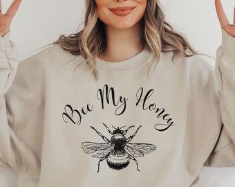 May include: Beige sweatshirt with the text "Bee My Honey" in a script font, and a detailed black and white illustration of a bee. The sweatshirt is paired with a beige beanie.