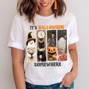 May include: White t-shirt with a Halloween-themed graphic. The design features a ghost, black cat, skeleton, and pumpkins, with the text "IT'S HALLOWEEN SOMEWHERE" in orange and black.