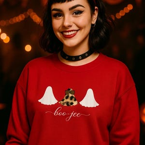 May include: A red Halloween-themed sweatshirt with a design featuring two white ghost figures flanking a leopard print bow and the cursive text "boo-jee". The sweatshirt is a festive garment for the spooky season.