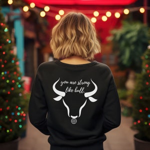 Puede incluir: Sudadera negra con un gráfico de toro blanco y el texto "you are strong like bull." El diseño presenta una cabeza de toro con cuernos y un elemento decorativo. La sudadera es de cuello redondo.