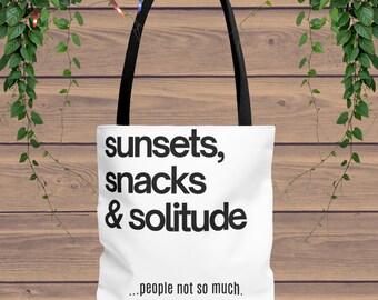 Może przedstawiać: Biała torba typu tote z czarnymi uchwytami. Torba ma napis "sunsets, snacks & solitude" w czarnym, pogrubionym kroju pisma. Poniżej znajduje się fraza "...people not so much." Torba jest umieszczona na drewnianym tle.