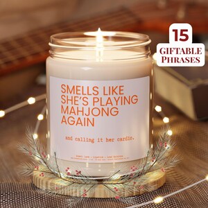 Puede incluir: Una vela encendida en un tarro de cristal con una etiqueta blanca. La etiqueta dice "Smells Like She's Playing Mahjong Again" en naranja. La vela está sobre un posavasos de madera con una ramita decorativa.