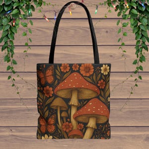 May include: Black tote bag featuring a whimsical mushroom and floral design. The bag showcases orange mushrooms with white spots, butterflies, and flowers against a dark background. It has black handles.