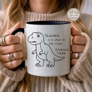 May include: White ceramic mug with a black interior and handle. The mug features a black and white cartoon dinosaur and the text "Teaching is a walk in the park... Jurassic Park."