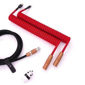 Puede incluir: Un cable rojo en espiral con dos conectores dorados y un cable negro con un conector negro. El cable rojo está en espiral y el cable negro está recto. Hay un pequeño objeto con forma de panda en la imagen.