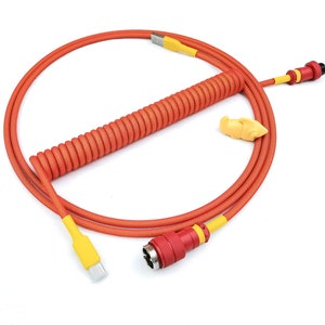 Puede incluir: Un cable naranja enrollado con extremos amarillos y un conector rojo. El cable está enrollado en círculo y tiene una pieza de plástico amarilla unida.