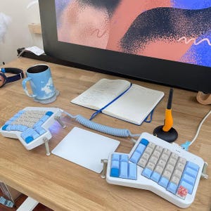 Könnte beinhalten: Eine ergonomische Tastatur in Hellblau und Weiß mit Spiralkabel, eine Tasse mit Wolkenmuster und ein Notizbuch auf einem Holztisch. Ein digitales Zeichentablett und ein Stift sind ebenfalls vorhanden.