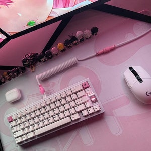 May include: A white and pink mechanical keyboard with a pink cable and a white mouse on a pink desk mat. There are several small figurines on the desk, and two computer monitors with anime images on the screen.