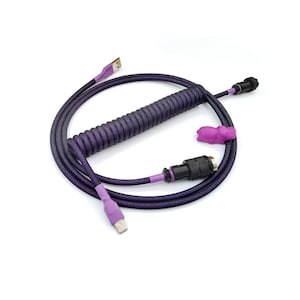 Pode incluir: Cabo USB roxo e preto enrolado, com um conector roxo correspondente e uma pequena seção enrolada. O cabo tem um conector USB-A em uma extremidade e um conector USB-C na outra. Uma abraçadeira roxa está presa.
