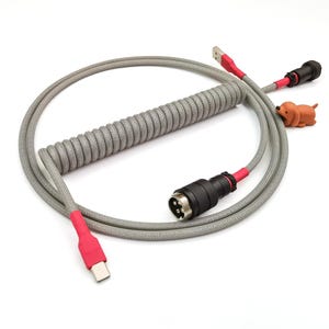 May include: A gray coiled cable with red and black connectors. The cable has a small orange rubber dog charm attached.