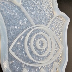 May include: A clear silicone mold shaped like a hamsa hand with an eye in the center. The mold is filled with clear glitter.