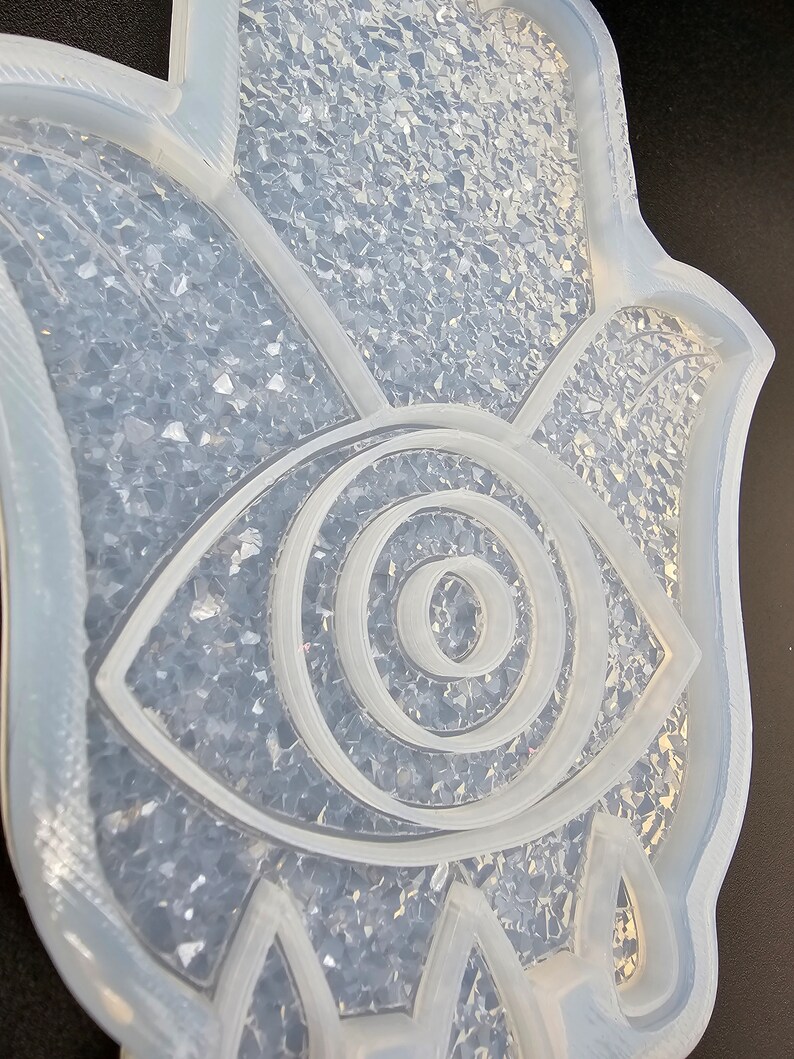 May include: A clear silicone mold shaped like a hamsa hand with an eye in the center. The mold is filled with clear glitter.