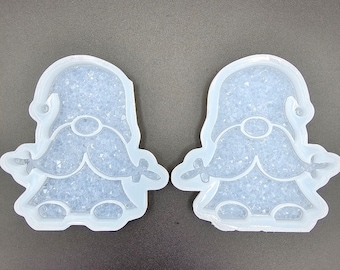 May include: Two clear, gnome-shaped molds filled with a sparkly, light-colored substance. The molds have a detailed outline of a gnome with a hat, beard, and arms. The molds are made of a flexible material and are on a dark gray surface.