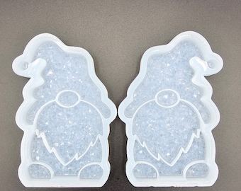 May include: Two clear gnome-shaped molds with a textured, glittery interior. The molds are made of a flexible material and are designed with a hat, beard, and body shape. The molds are for crafting and DIY projects.