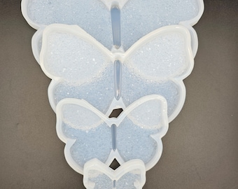 May include: Four clear, butterfly-shaped silicone molds of varying sizes are stacked on a dark surface. The molds have a textured interior and are designed for crafting or resin art projects. The largest mold is at the top, with the smallest at the bottom.