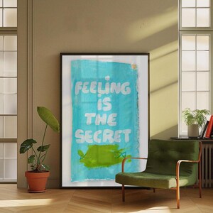 Può includere: Grande opera d'arte incorniciata con la scritta "FEELING IS THE SECRET" in bianco su sfondo turchese. Una forma astratta verde in basso. L'opera è esposta in una stanza con una sedia verde e una pianta in vaso.