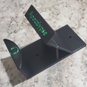 May include: Black 3D printed wall mount with a green inscription that reads "Apocalypse". The mount has a curved section with a green "C" symbol.