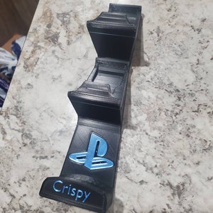 Op de afbeelding: Een zwarte 3D-geprinte PlayStation-controllerstandaard met een blauw PlayStation-logo en het woord "Crispy" gedrukt op de voorkant.