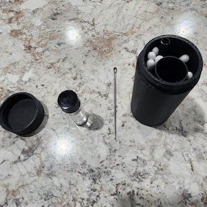 May include: A black cylindrical container with a lid, a small glass vial with a black cap, and a metal tool are arranged on a speckled surface. The container holds several white objects. The items are positioned for easy access.