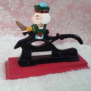 May include: A black cast iron nutcracker with a red wooden base. A painted figure of a nutcracker is riding on top of the nutcracker.