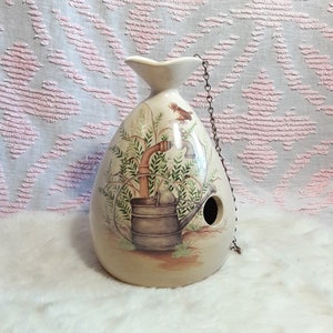 以下が含まれることがあります： 鳥の形をしたセラミック製のバードハウス。ジョウロの形をしていて、鳥が注ぎ口にとまっている。バードハウスは、茶色のジョウロ、緑の葉、茶色の鳥が描かれている。バードハウスには、鳥が出入りできる穴と、吊り下げ用のチェーンが付いている。