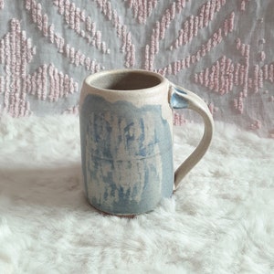 Puede incluir: Una taza de cerámica azul y blanca con asa. La taza tiene un aspecto rústico y hecho a mano y está esmaltada en un color azul claro con detalles blancos.