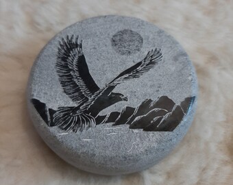 Può includere: Una pietra rotonda grigia con un'immagine scolpita di un'aquila in volo sullo sfondo di montagne e luna piena. L'aquila è nera e la luna è bianca.