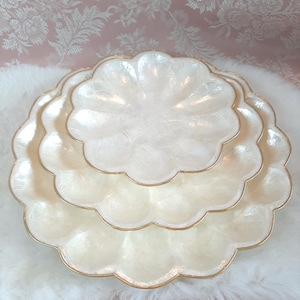 May include: Three off-white, scalloped serving dishes with a pearlescent finish and gold trim. The dishes are stacked on a white, fluffy surface. The background features a pink floral pattern.